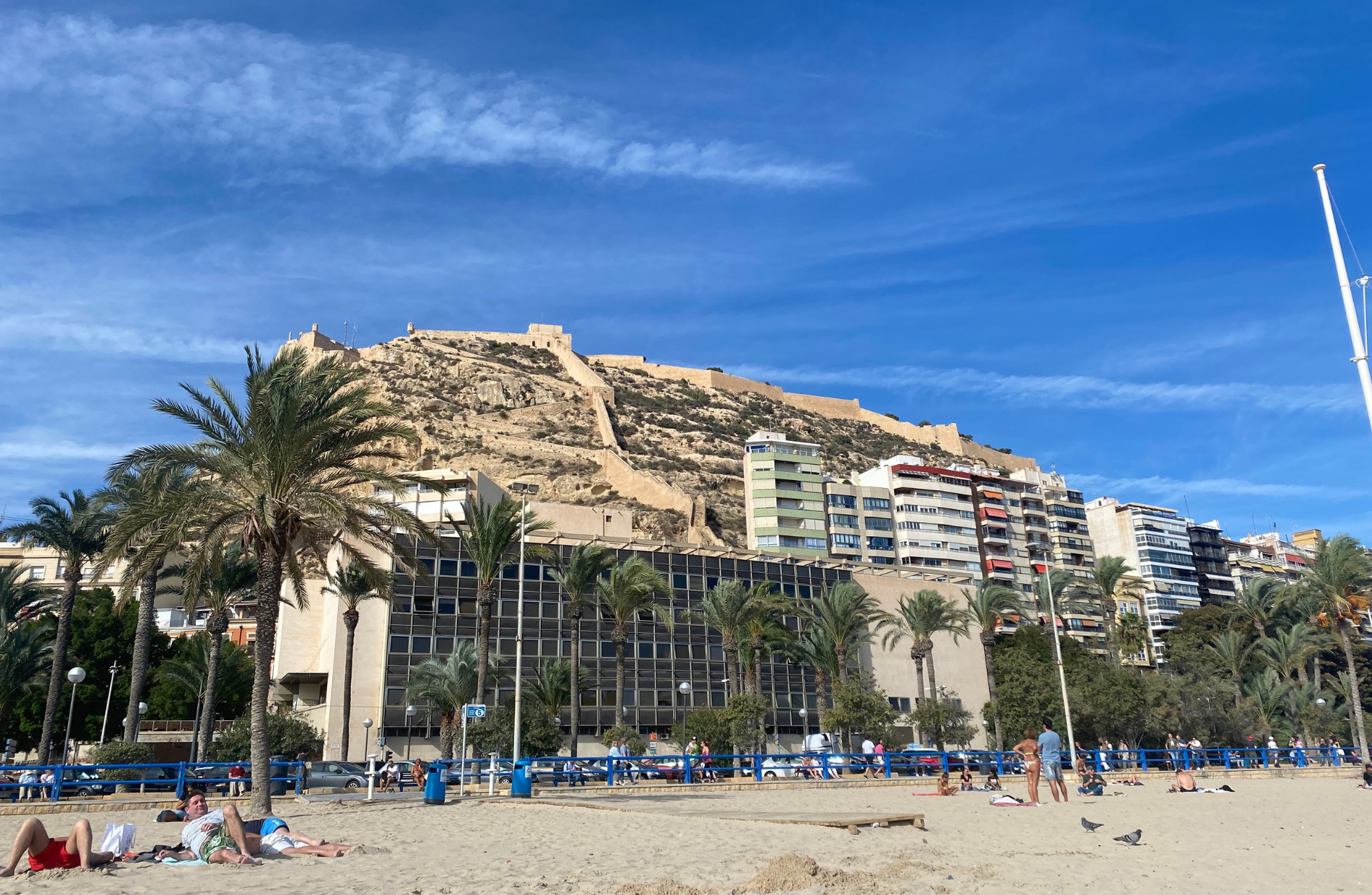 view from the beach up with modern buildings in front of a mountain with medieval castle-like structures on the mountaintop