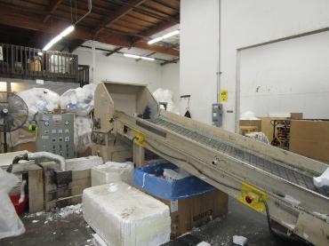 A large machine with a conveyor belt. Surrounded by Styrofoam products.