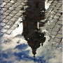 A building spire, clouds and and sky reflected in a puddle on a cobblestone street.