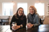 photo of Coya Paz Brownrigg and Chloe Johnston sitting at a wooden table and holding coffee mugs, smiling at the camera.