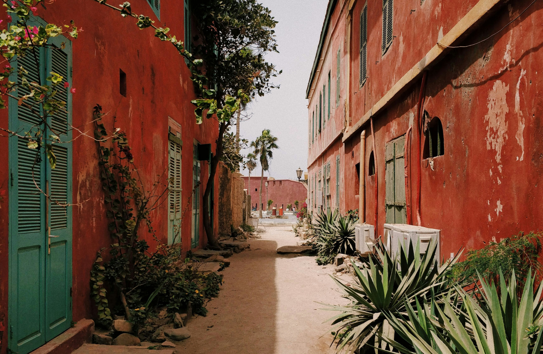 a street in along two red buildings on either side and a hot environment with tropical plants around