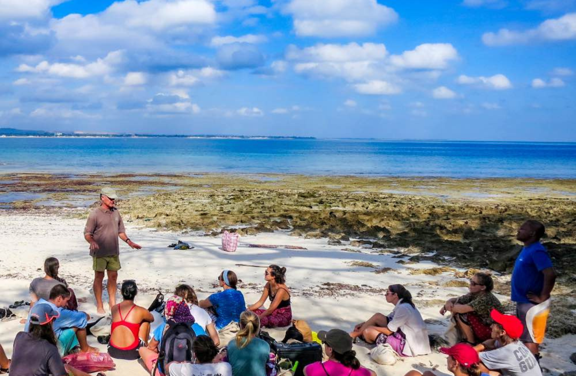 students sitting on a beach listening to a standing lecturer