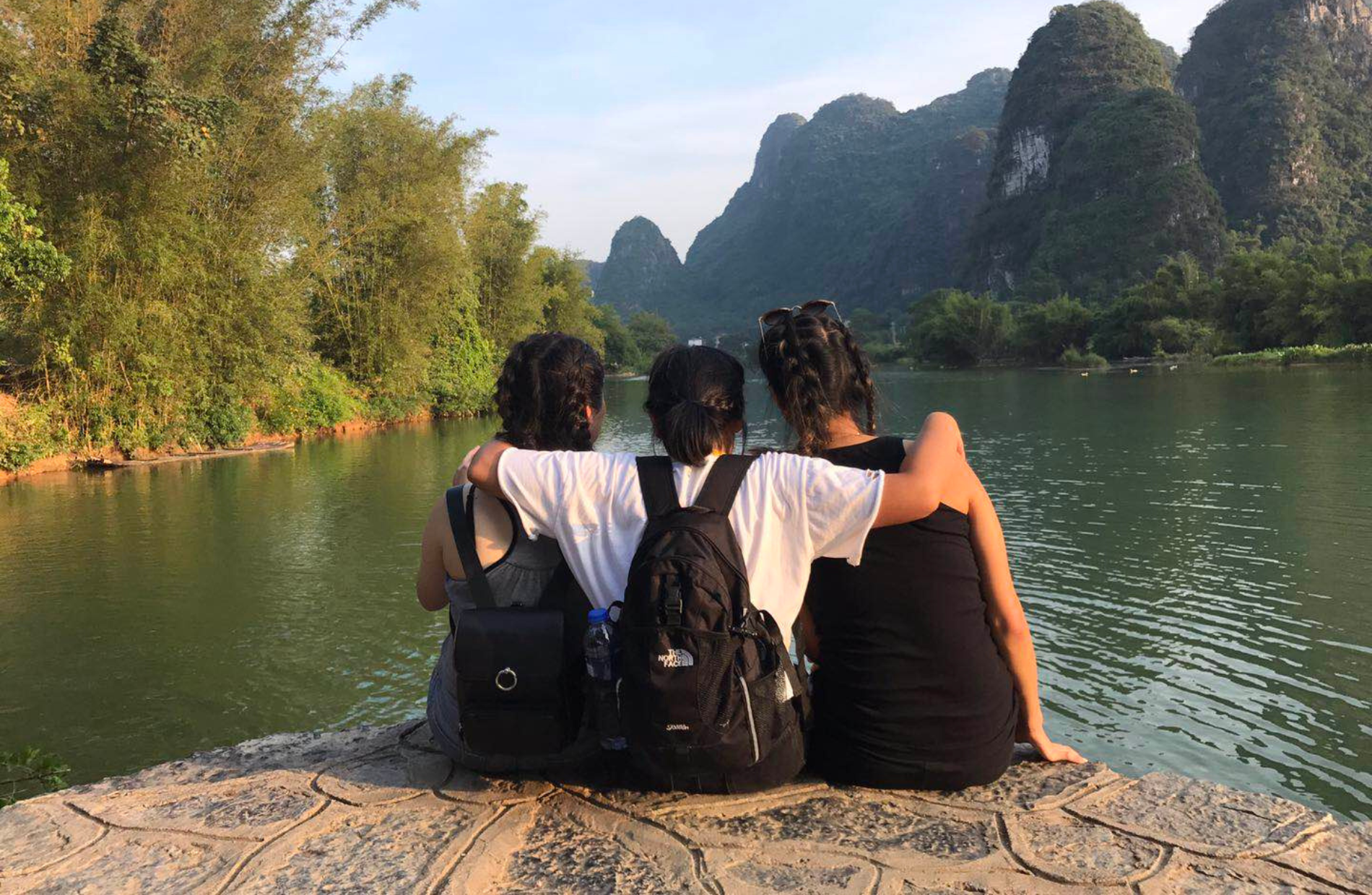 the backs of three women sitting along a mass of water with mountains in the background