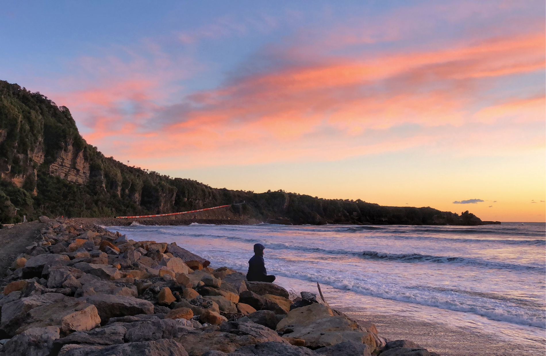 Woman looking out on the ocean while sitting on a beach at a sunset at Punakaiki Beach