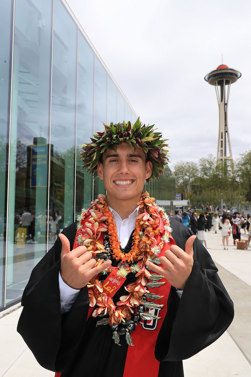 SU graduate with flower leis and crown smiles and gives two shaka signs in front of the Seattle Space Needle.