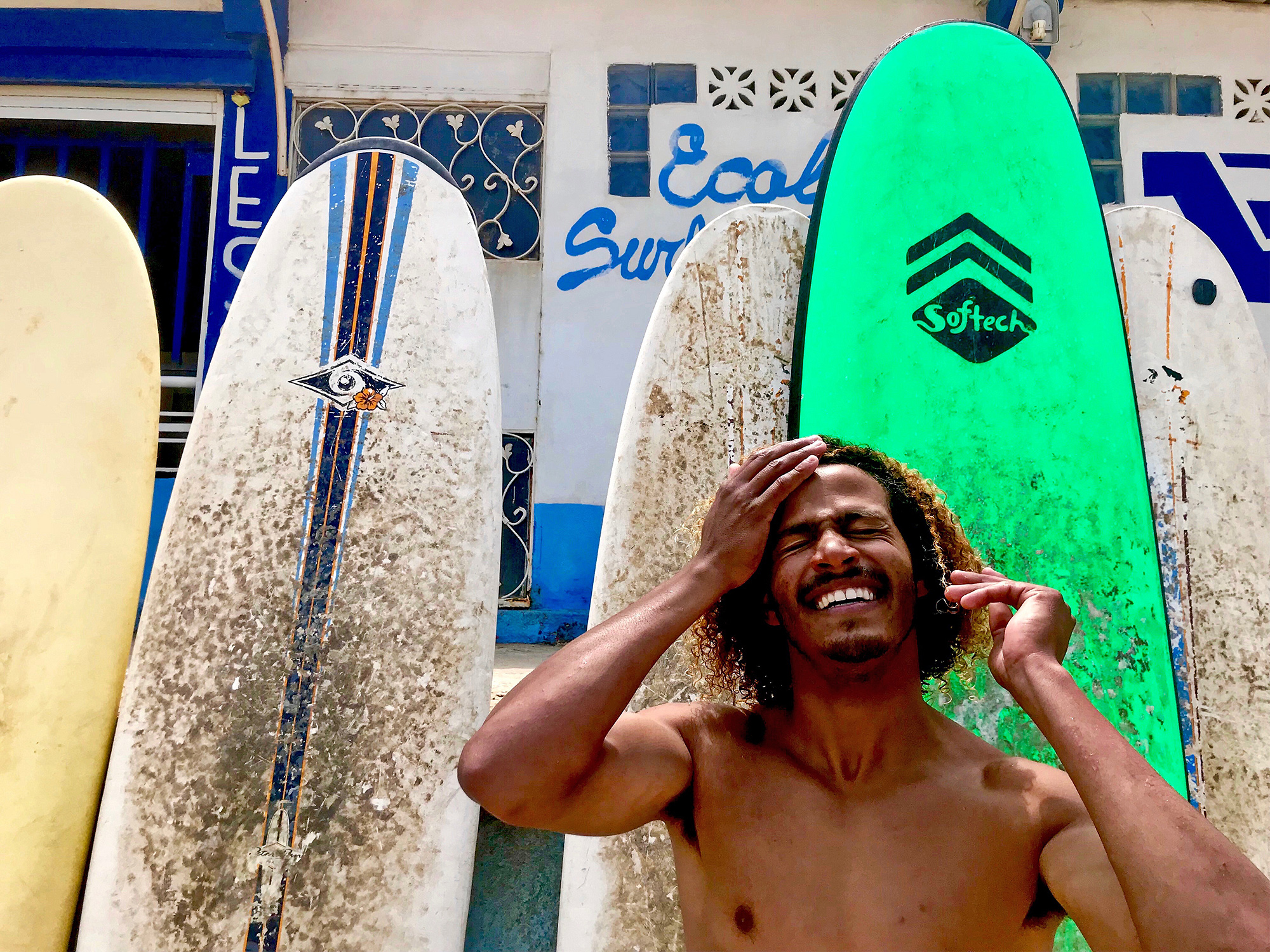 a shirtless man smiles in front of several surfboards leaning against a tiled wall