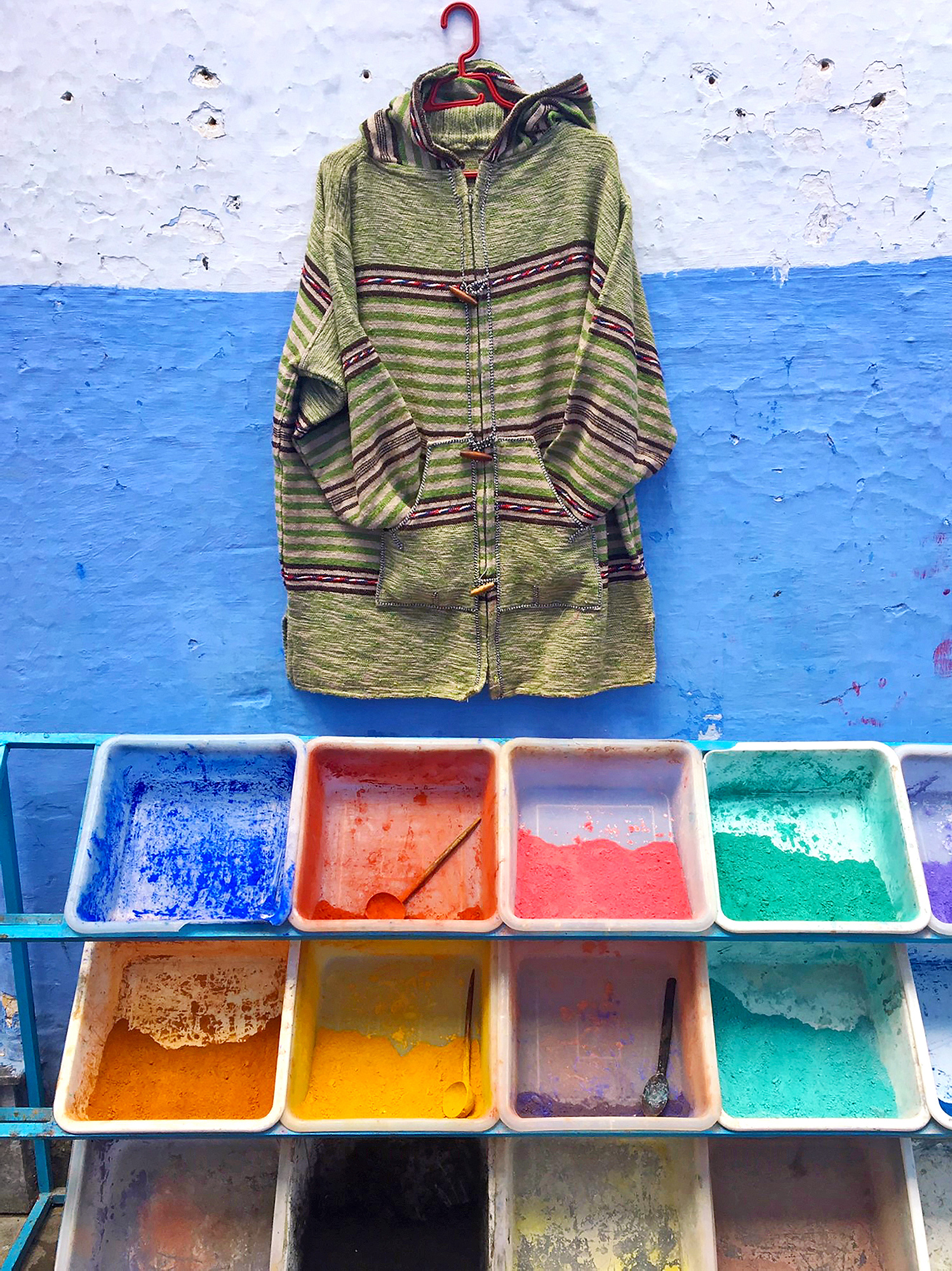 a zippered hooded jacket hangs on a wall painted white and blue above open bins of colored pigment