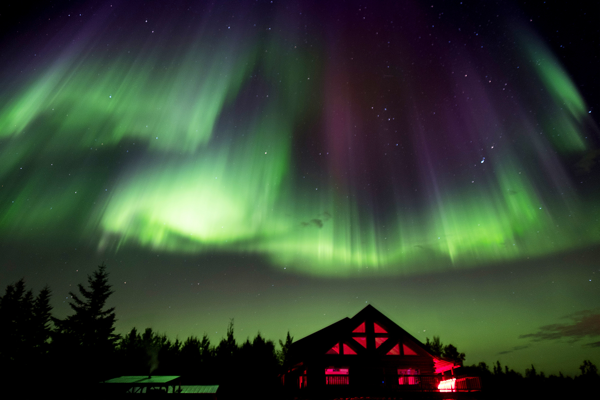 a cabin glows a warm red beneath the green and purple Aurora Borealis lights in a starry night sky