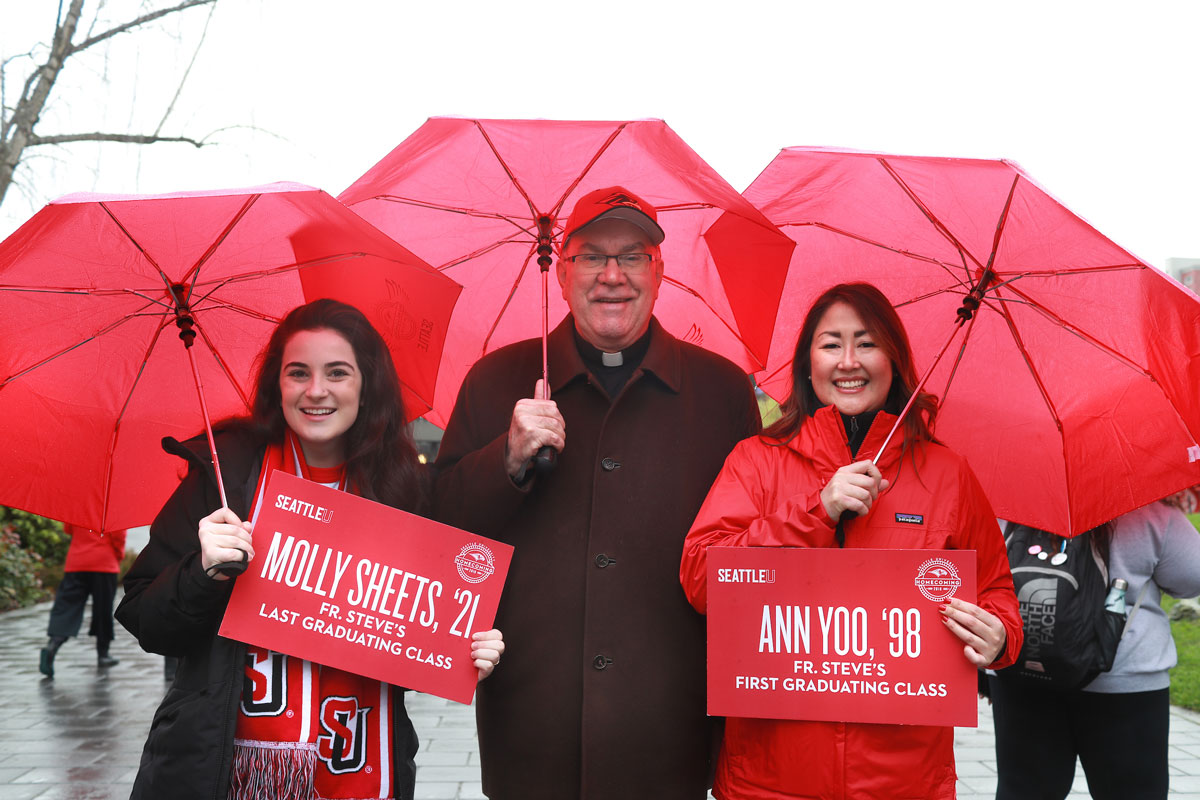 Father Steve is flanked by two alumni, all three holding umbrellas.