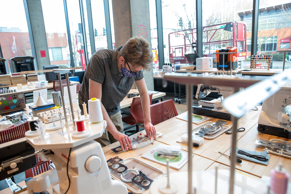 a student at a large wooden table working on a hands-on project amidst machines in a daylight-fllled makerspace